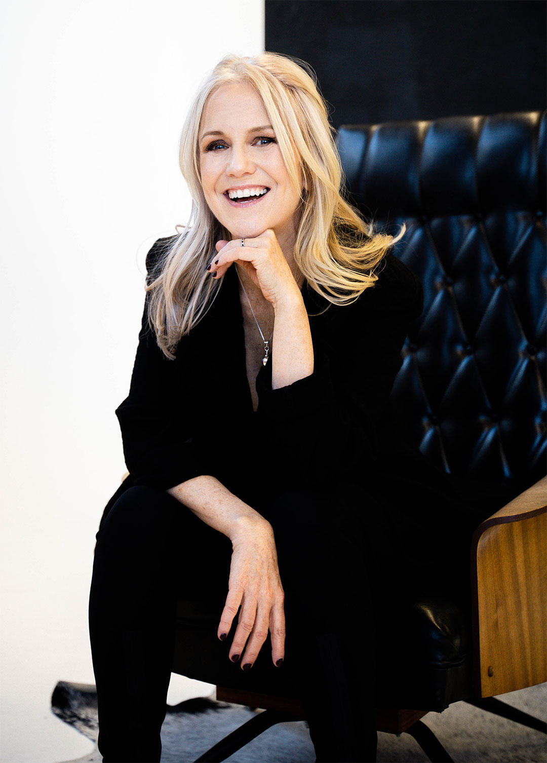 Nina Rolle smiling and wearing a black suit while sitting in a black leather armchair